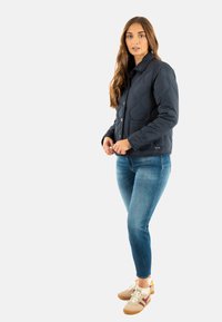 Giovane donna con capelli lunghi che indossa una giacca trapuntata scura, jeans blu e sneakers beige, in piedi contro uno sfondo semplice.