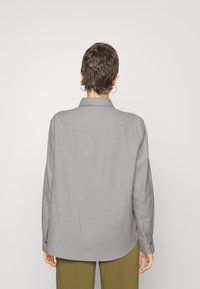 Maglietta grigia a maniche lunghe con colletto, dotata di una trama liscia, taglio dritto e polsini abbottonati, indossata con pantaloni verde oliva.