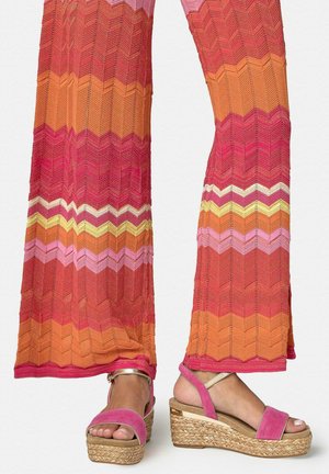 Piernas con pantalones de pierna ancha con patrón de zigzag colorido en rojo, naranja, rosa y amarillo, con sandalias de plataforma rosa con tiras en el tobillo.