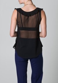 Blusa nera trasparente con colletto a ruches plissettato, design senza maniche e orlo curvo; abbinata a pantaloni blu per un look elegante.