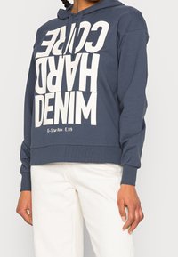 Marinblå hoodie med stor vit text "HARD CORE DENIM" och logotyp, med snörning i huvan och ribbade ärmslut, bärs över ljusa byxor.