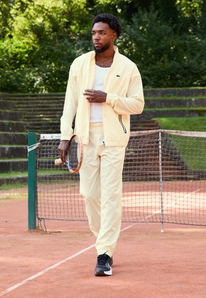 Gaiši dzeltena treniņu uzvalks ar rāvējslēdzēja jakas, elastīgām piedurknēm, balta krekla apakšā, melnas sporta kedas, tur tennis izsistāju uz māla laukuma.