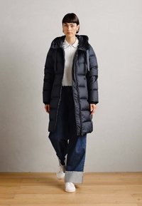 Lang, puffy svart jakke med hette, glidelås foran og ribbestrikkede mansjetter. Kombinert med brede bukser i blå denim og hvite joggesko. Glatt tekstur.