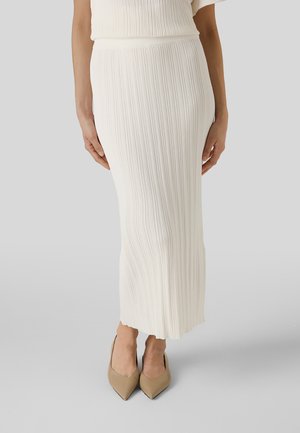 Femme portant une jupe midi en maille côtelée blanche et des talons beiges à bout pointu, debout devant un fond gris clair uni.