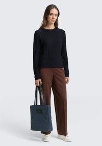 Marineblauer gerippter Pullover kombiniert mit braunen weiten Hosen; hält eine dunkelblaue Tote-Bag mit langen Trageriemen; weiße Sneakers.