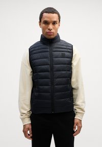 Μαύρο γιλέκο με κουμπί, με κρεμαστή ραφή, ψηλό γιακά και κλείσιμο με φερμουάρ, τοποθετημένο πάνω από ένα ανοιχτό κρεμ πουκάμισο μακρυμάνικο και μαύρα παντελόνια.