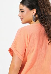 Top di colore pesca con maniche corte arrotolate, caratterizzato da una vestibilità ampia e una finitura in tessuto leggermente stropicciato. Orecchini metallici di grandi dimensioni.
