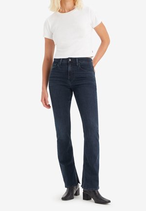 Femme portant un t-shirt blanc court, un jean taille haute bleu foncé avec des fentes latérales, et des bottines noires à talons, debout avec une main sur la hanche.