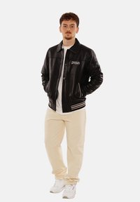 Schwarze Lederbomberjacke mit weißem Logo, gestreiften Bündchen, kombiniert mit cremefarbenen Hosen und weißen Sneakern, vor einem schlichten Hintergrund stehend.