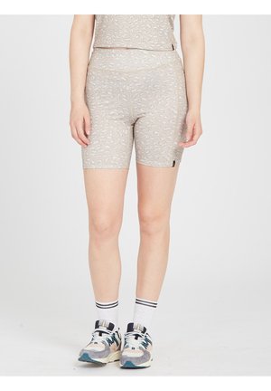 Femme portant un short beige à motifs, des chaussettes blanches à rayures noires et des chaussures de sport multicolores, debout devant un fond uni.