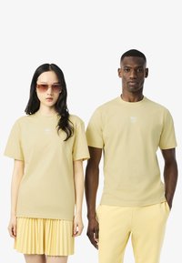 T-shirt in cotone di colore giallo chiaro, con scollo rotondo e logo ricamato sul petto. La modella abbina con una gonna plissettata e pantaloni coordinati.