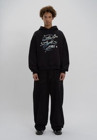 Zwarte capuchontrui met een grafische print, gedragen met losvallende zwarte broek en zwarte schoenen. Eenvoudige, gestructureerde stof.