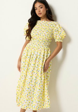 Vrouw met lang donker haar in een witte midijurk met gele citroen- en groene bladprint, geplooide taille en opgeblazen korte mouwen.