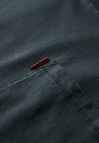 Mørkegrøn stof med en glat tekstur, der har et rødt mærke ved lommen med teksten "SUPERDRY." Enkelt design med rene kanter.