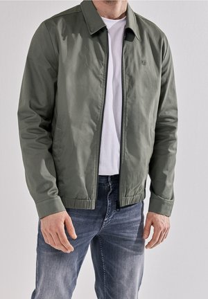 Mand iført en grøn lynlåstrøje over en hvid T-shirt og blå jeans, stående mod en ensfarvet lys baggrund.