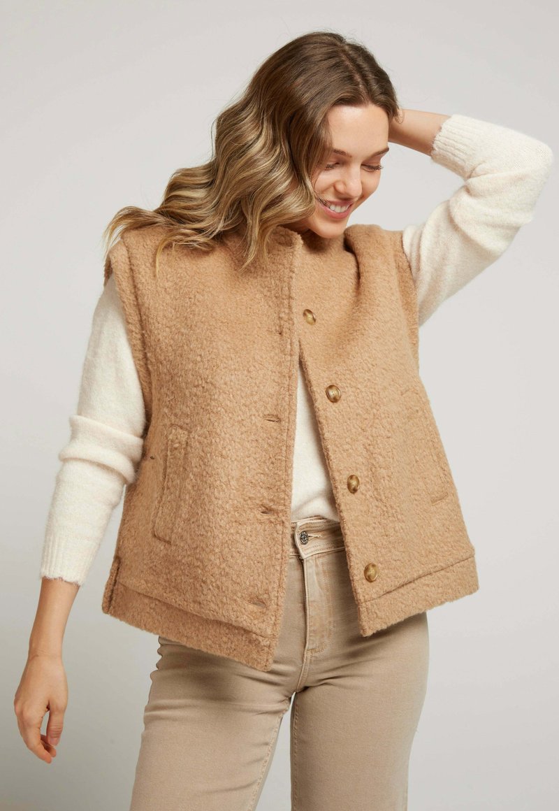 Gilet en laine beige avec une surface texturée, col rond et fermeture à boutons sur le devant. Il possède deux poches latérales et est porté par-dessus un pull crème.