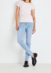 Světle růžové bavlněné tričko s malým logem, v kombinaci se světle modrými slim-fit džíny a černými mokasíny, na bílém pozadí.