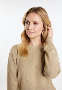 Beige Strickpullover mit einer weichen, strukturierten Oberfläche. Verfügt über einen runden Halsausschnitt und lange Ärmel. Das Model hat lange, lockige Haare und trägt goldene Ohrringe.