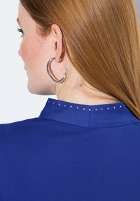 Modrá blůza s vysokým límcem, zdobená stříbrnými cvočky podél výstřihu. Modelka má na sobě velké stříbrné náušnice ve tvaru kruhu. Hladký materiál, rovný střih.