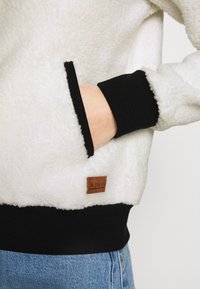 Biały polarowy sweter z czarnymi ściągaczami na mankietach i dole, z kieszenią z przodu oraz brązową skórzaną naszywką z logo Roxy na dolnej części przodu.