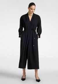 Musta haalari syvällä V-aukolla, pitkillä hihoilla, vyötetty vyötärö ja leveät lyhyet lahkeet; sisältää sivutaskut ja sileän kankaan.