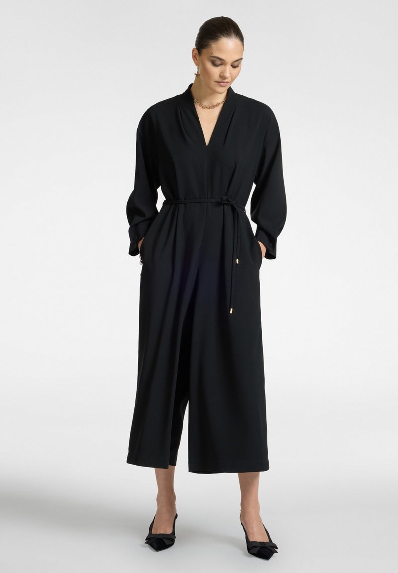 Musta haalari syvällä V-aukolla, pitkillä hihoilla, vyötetty vyötärö ja leveät lyhyet lahkeet; sisältää sivutaskut ja sileän kankaan.