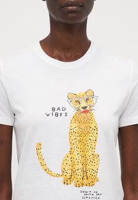 Balta kokvilnas T-krekls ar dzeltenu leopardu ilustrāciju ar melnajiem plankumiem, kas nēsā brilles, un tekstu "Slikti vibrācijas" un "Neiet kopā ar manu lūpu krāsu."