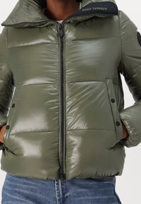 Giaccone imbottito verde oliva lucido, caratterizzato da collo alto, chiusura con zip e bottoni automatici neri sulle tasche, con texture trapuntata.