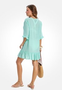 Mintgroene jurk met driekwartmouwen en gerimpelde zoom. Lichtgewicht stof, ontspannen pasvorm. Model draagt een strooien handtas.