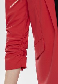 Blazer rojo con una textura suave, que presenta un detalle fruncido en la manga, un bolsillo y un ajuste estructurado.