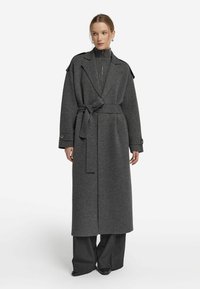 Szary wełniany płaszcz trench z paskiem, z szerokim kołnierzem, zapięciem na guziki i luźnym krojem. Stylizowany na sweter z prążkowanym golfem i luźnymi spodniami.