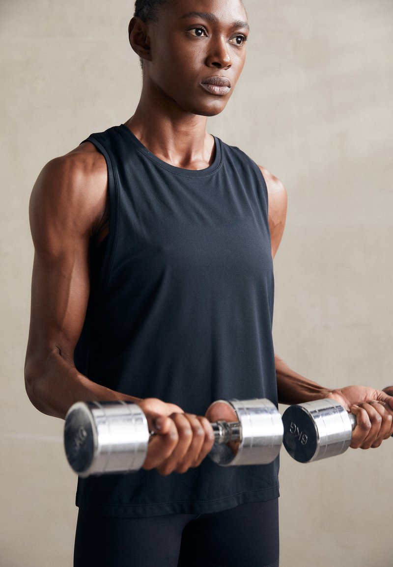 Zwarte mouwloze sporttop; gespierde armen die zilveren dumbbells vasthouden; gladde stof; focus op kracht en fitness. Neutrale achtergrond.