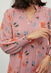 Femme portant une robe longue à manches longues rose à motifs floraux avec des poignets smockés et un décolleté en V, ajustant une manche avec une main.