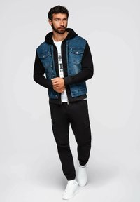 Giubbotto di denim con maniche nere, abbinato a una t-shirt bianca con grafica e pantaloni jogger neri, completato da sneakers bianche. Look casual.