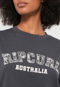 Sötétszürke póló bordázott, környakas kivitelben. A "RIPCURL" felirat virágmintás betűtípussal, alatta az "AUSTRALIA" felirat tiszta fehér színben. Puha pamut anyagból.