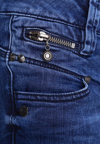 Los jeans de denim azul cuentan con un bolsillo con cremallera y herrajes de metal, dos botones visibles y una pequeña etiqueta con el logotipo. La tela tiene un aspecto ligeramente texturizado.