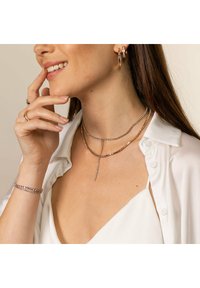 Schmuck aus Roségold mit geschichteten glitzernden Ketten im Lariat-Stil. Modell trägt Creolen und Ringe, gestylt mit einer weißen Bluse.