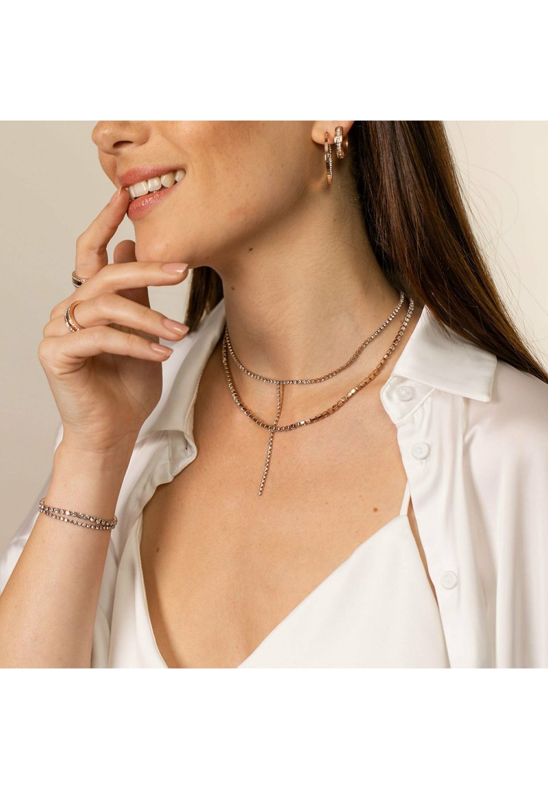 Schmuck aus Roségold mit geschichteten glitzernden Ketten im Lariat-Stil. Modell trägt Creolen und Ringe, gestylt mit einer weißen Bluse.