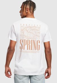 Hvid bomulds t-shirt med et tropisk bladdesign og styliseret tekst "TROPISK FORÅR" i lys brun på ryggen. Korte ærmer.