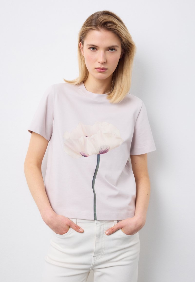 Junge Frau mit blondem Haar, die ein hellviolettes T-Shirt mit großem weißem Blumenmuster und weiße Hose trägt, Hände in den Taschen.