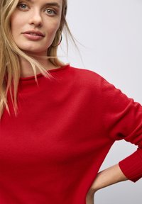 Maglione rosso con colletto e polsini a coste, realizzato in un morbido tessuto a maglia. Il design presenta una vestibilità rilassata e spalle leggermente abbattute.