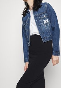 Jeansjacka i medelblått med en kortad design, metallknappar och en CK-logotyp patch på vänster bröst. Tillsammans med en svart kjol.