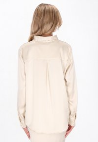 Beige satinbluse med bagpåsyet bærestykke og plisseringer, lange ærmer med knapmanchetter og klassisk kravedesign. Glat tekstur.