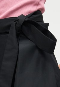 Μαύρη υφασμάτινη ζώνη δεμένη σε φιόγκο γύρω από τη μέση πάνω από μια ψηλόμεση μαύρη φούστα, συνδυασμένη με ροζ πλεκτό τοπ.