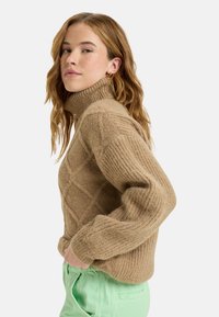 Strickpullover mit Rollkragen in Beige, mit einem strukturierten Diamantmuster und gerippten Bündchen; kombiniert mit mintgrünen Hosen.