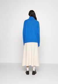 Persona con lunghi capelli scuri indossa un maglione a collo alto blu brillante, una gonna midi color crema, calze bianche e scarpe nere, in piedi di spalle.