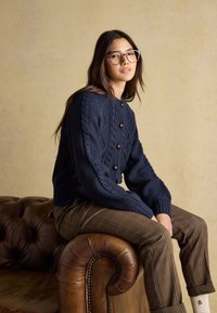 Ung kvinde med briller iført en navy kabelstrikket cardigan og ternede bukser, siddende på armlænet af en brun lædersofa.