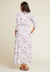 Lang blomstret kjole i lett stoff, med puffede ermer, midjebånd og et mønster av brune og kremfargede blomster på hvit bakgrunn.