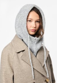 Γκρι πλεκτός σκούφος με μακριές κορδέλες, φορεμένος πάνω από ένα ανοιχτό καφέ παλτό με υφητό ύφασμα και διπλές κουμπότρυπες.