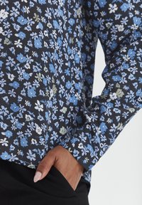 Blommig långärmad blus med blå, vita och gröna blommor på en svart bakgrund. Mjuk tygkvalitet med en avslappnad passform och manschetter på ärmarna.